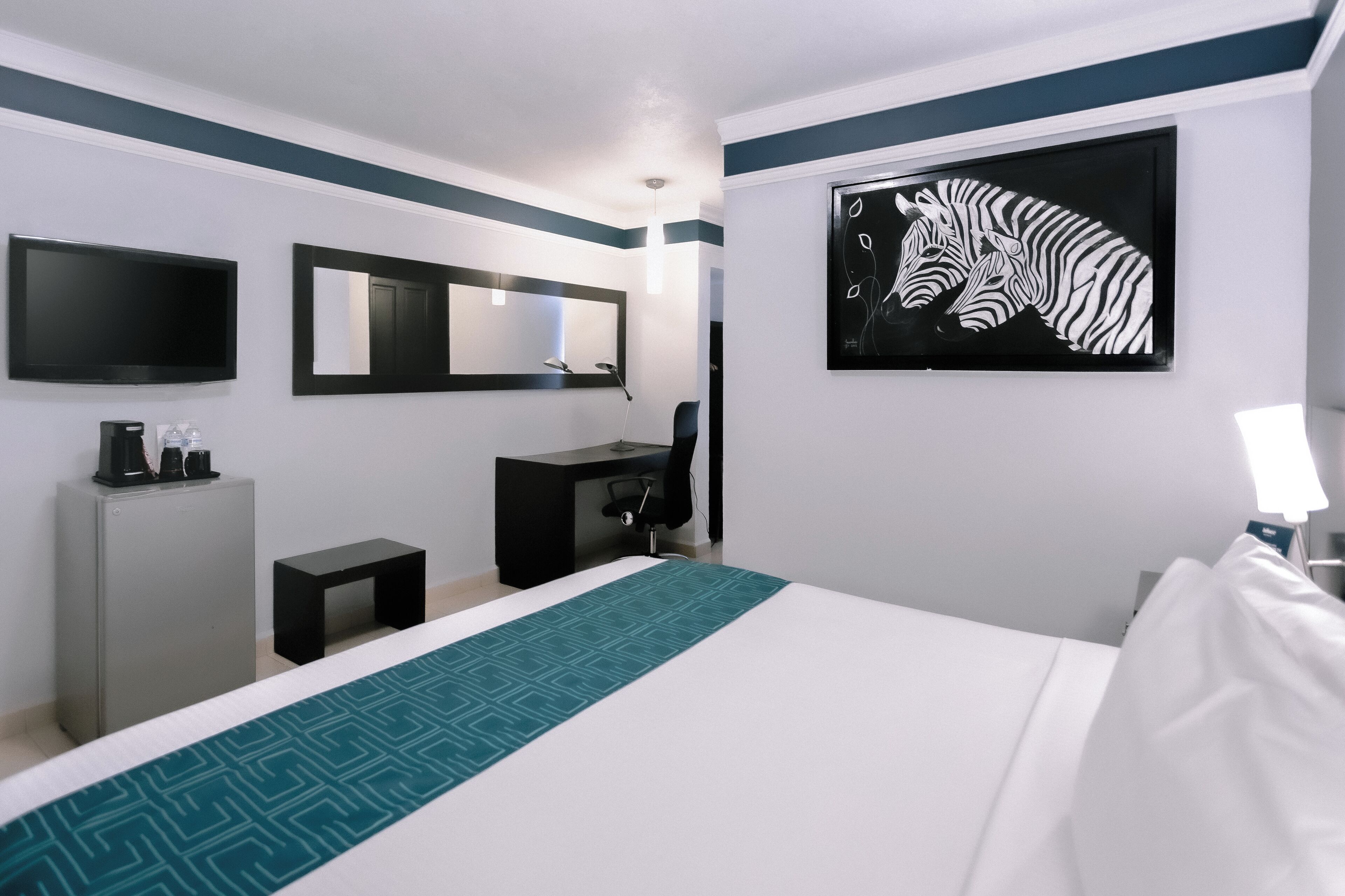 A hotel room with a bed, a desk, a chair, a mirror, a television, and a zebra artwork on the wall.