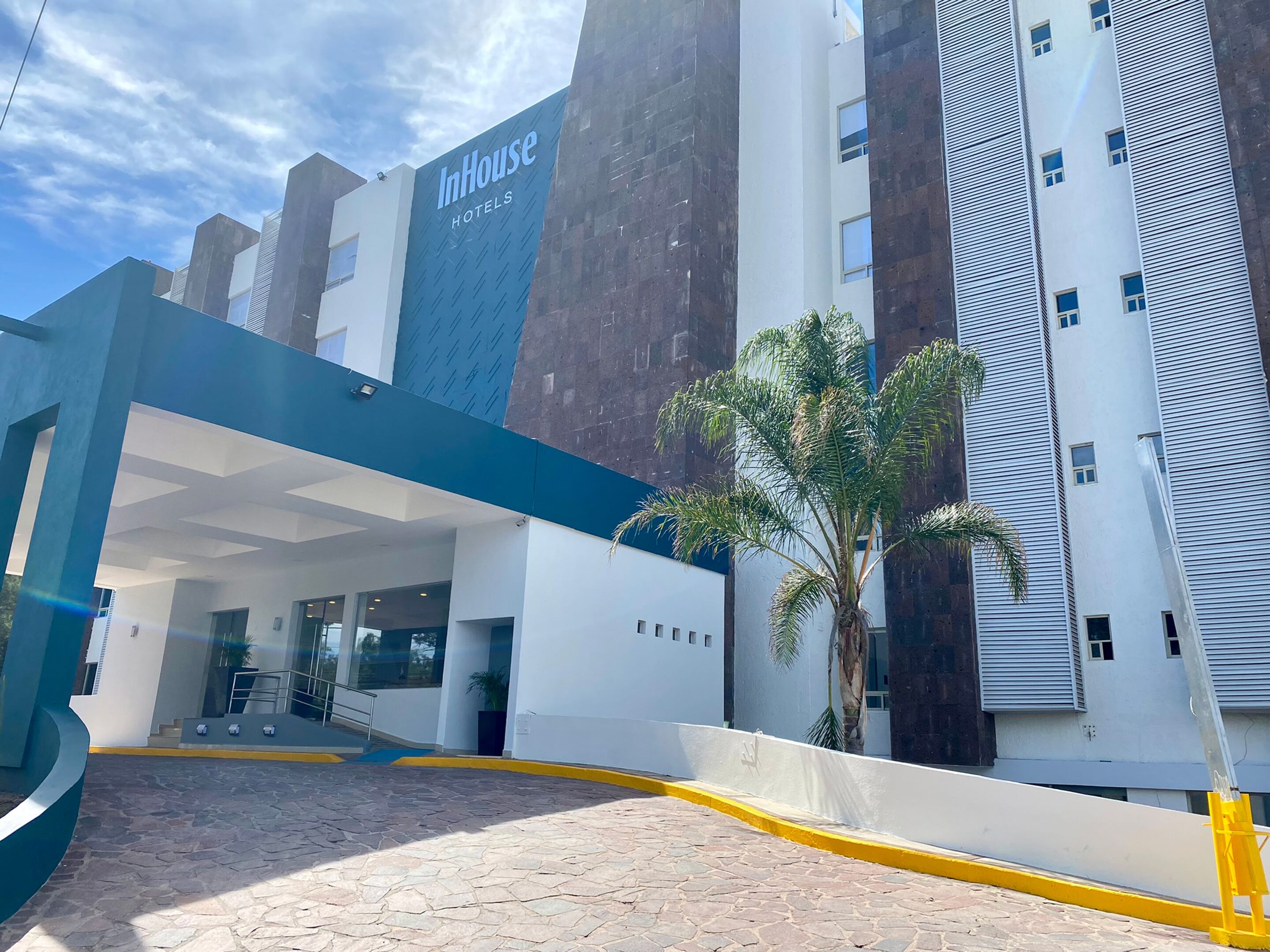 Uma entrada de hotel moderna com fachada azul e branca, uma palmeira e céu claro.