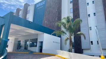 Uma entrada de hotel moderna com fachada azul e branca, uma palmeira e céu claro.