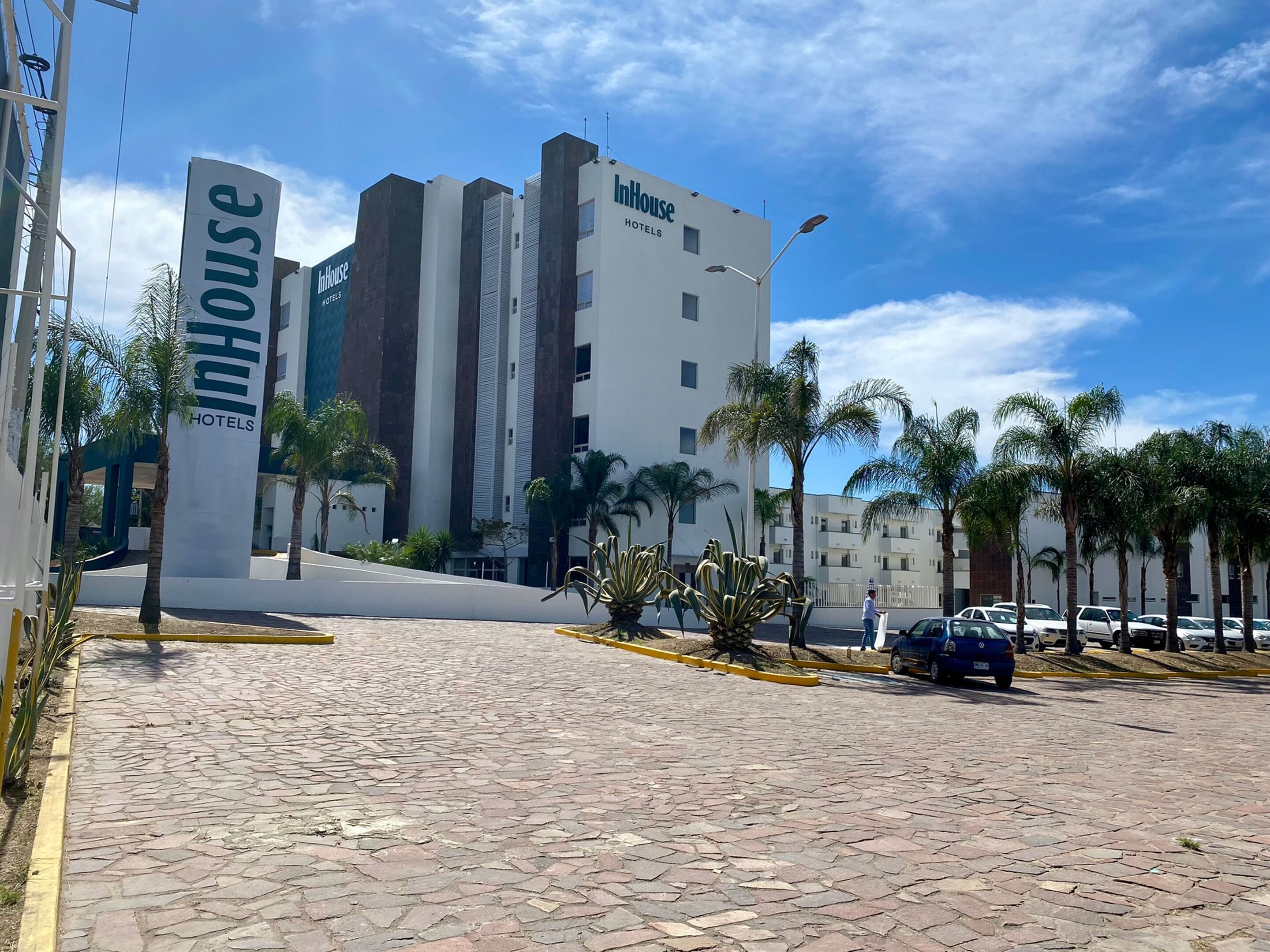 Entrada de un hotel con un letrero que dice "InHouse Hotels" y un camino pavimentado con palmeras.