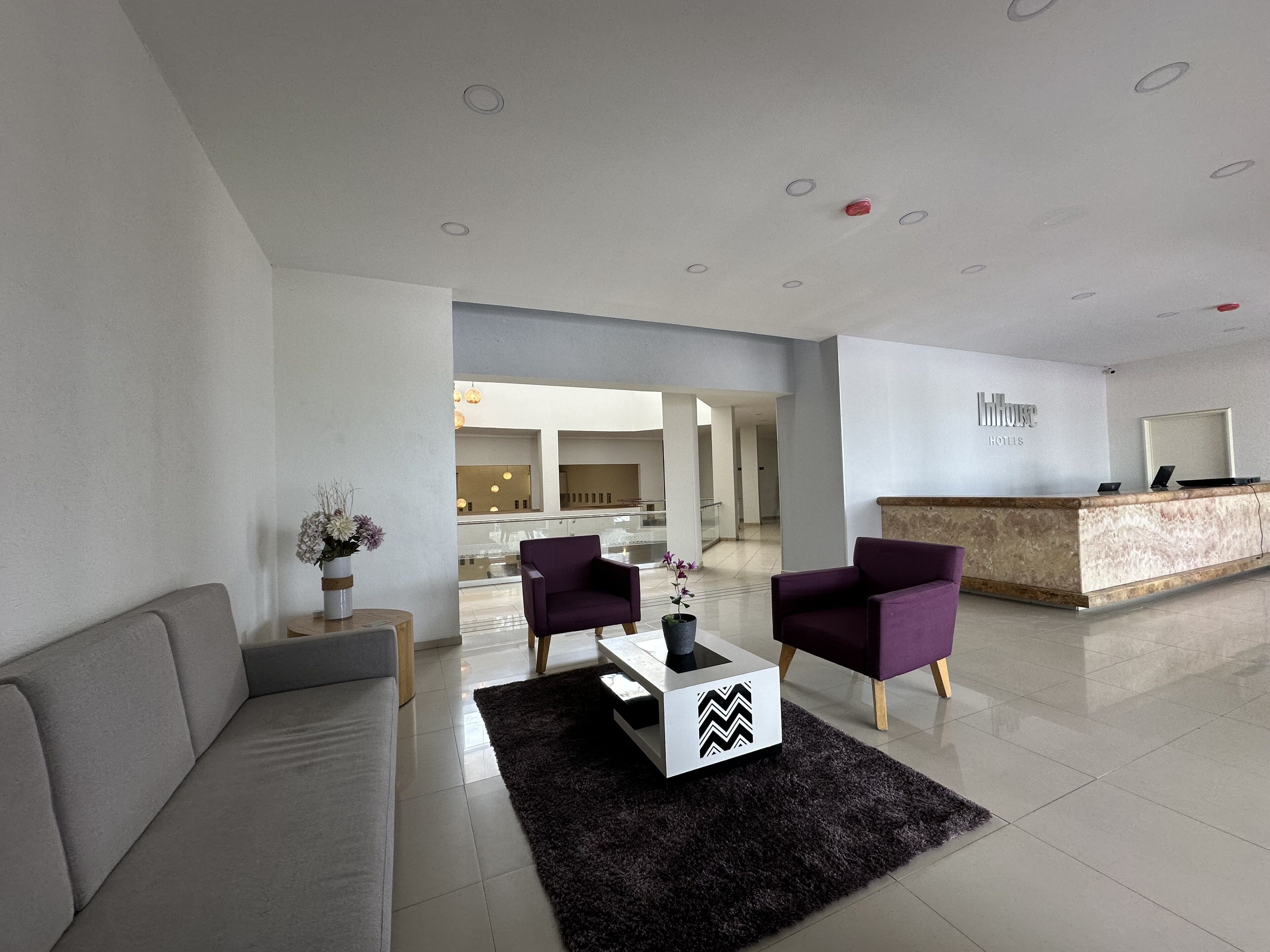 A modern hotel lobby with a reception desk, a grey sofa, two purple armchairs, and a black and white coffee table.