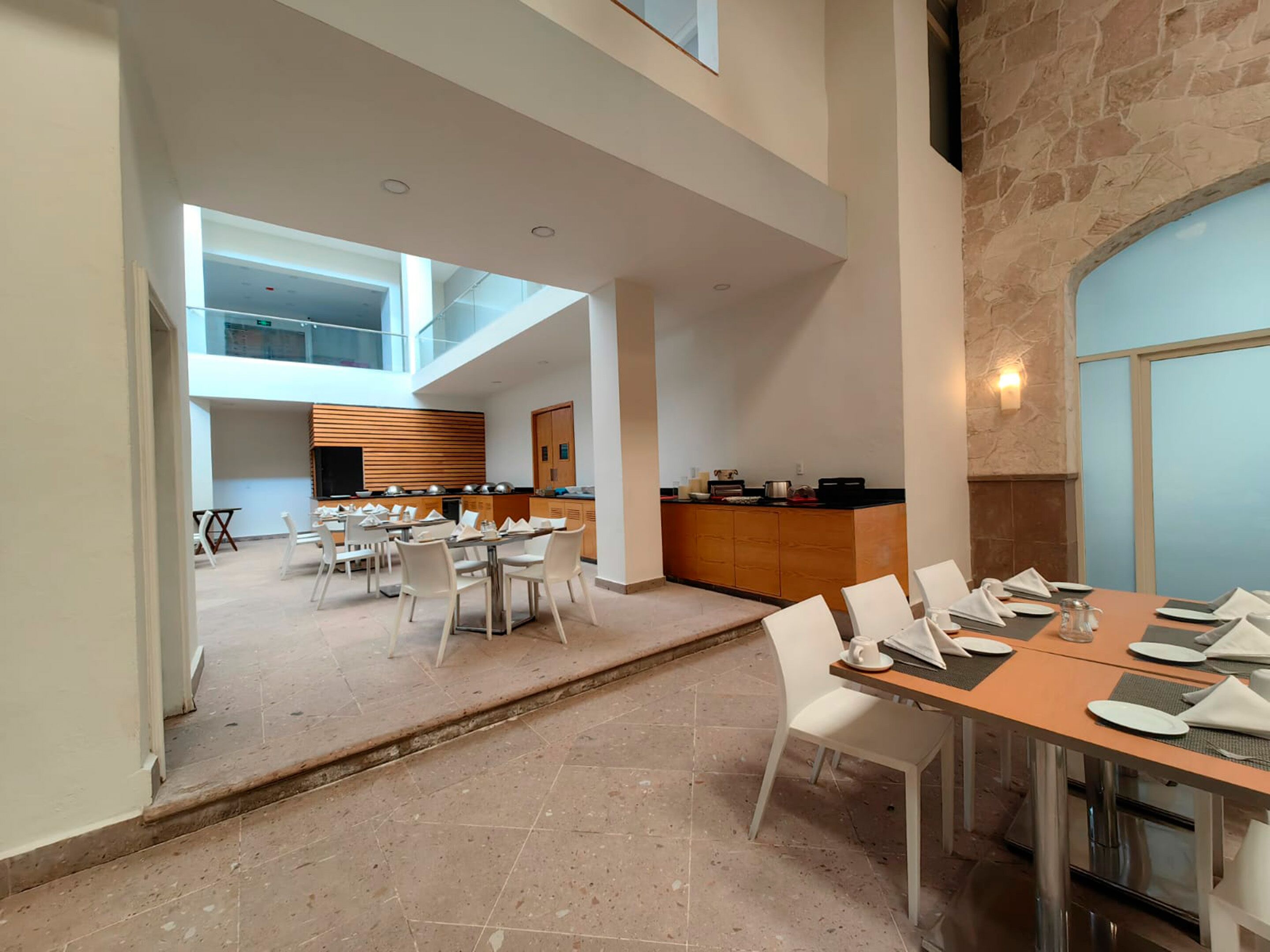 A spacious dining area with multiple tables and chairs, a kitchen in the background, and a stone wall.
