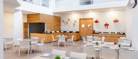 A modern hotel lobby with a central dining area, a reception desk, and a staircase leading to an upper level.