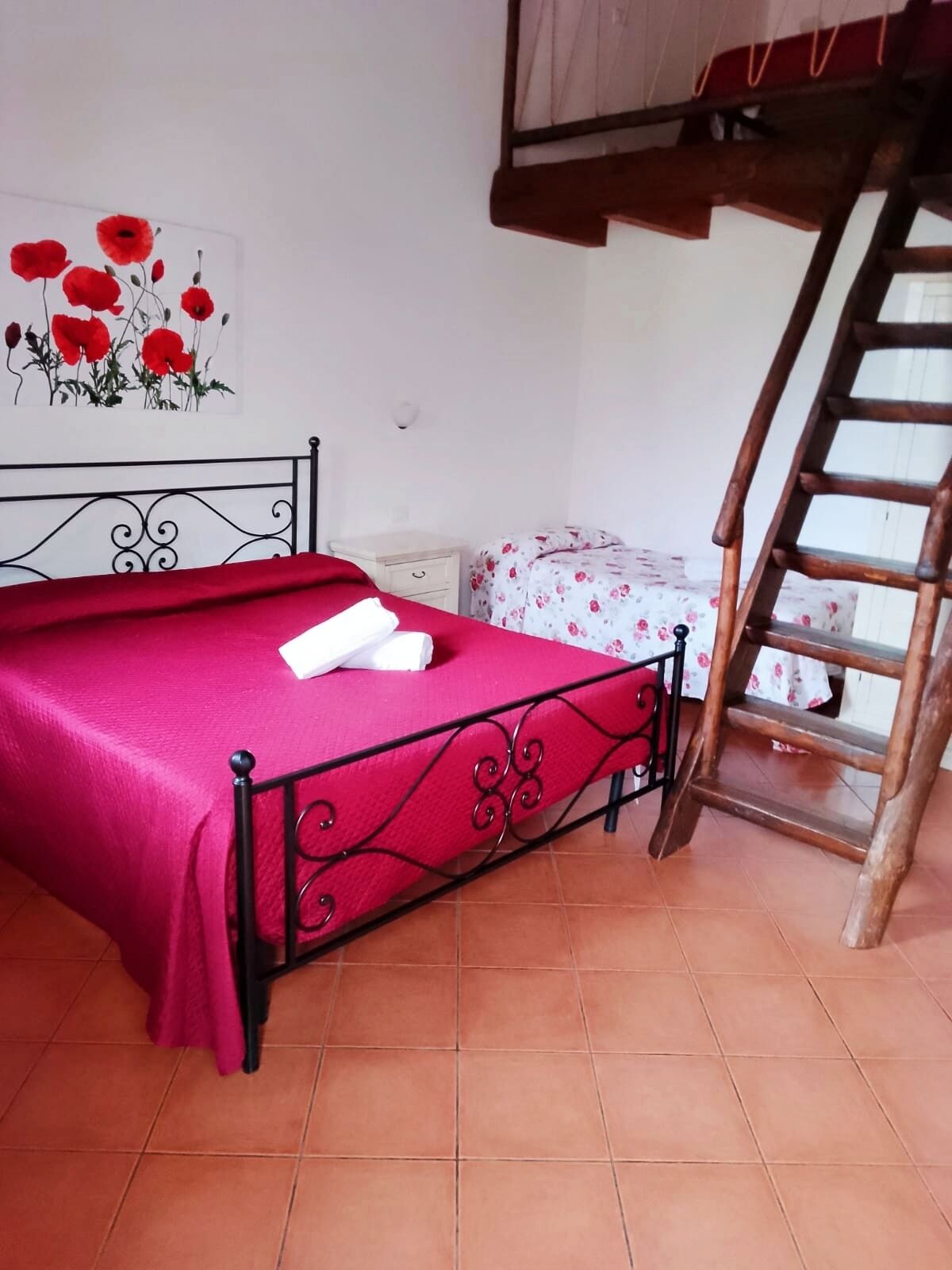 Een slaapkamer met een metalen bedframe, een rode bedspreider, een witte nachtkastje, een fauteuil met bloemenmotief en een houten ladder.