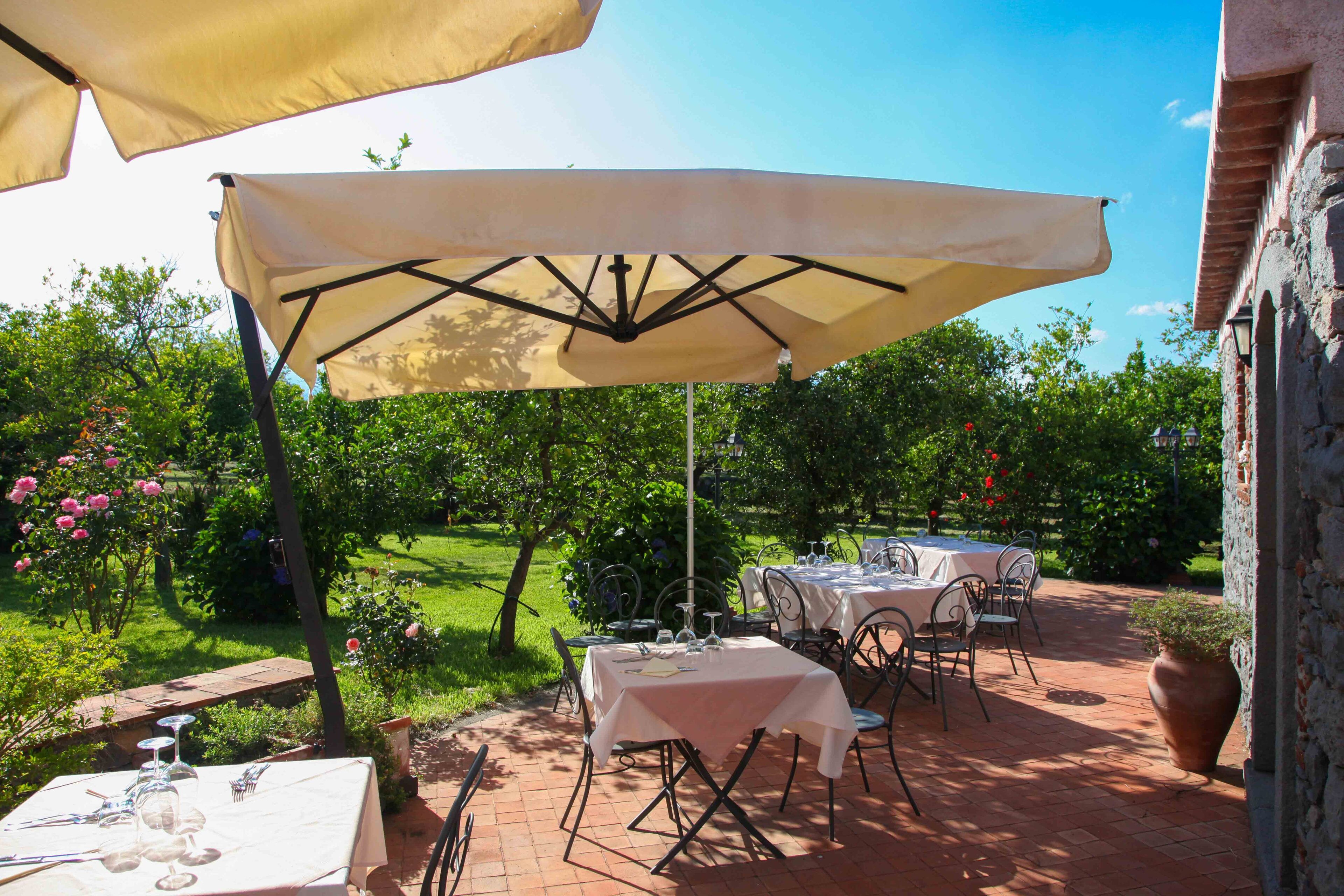 An outdoor patio with tables and chairs, covered by a large umbrella, set against a backdrop of greenery and a brick wall.