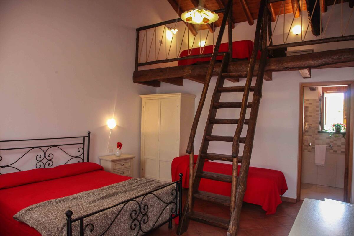 A bedroom with a bunk bed, a wooden ladder, a nightstand, a white wardrobe, and a bathroom visible through an open door.