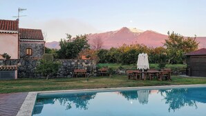 A swimming pool with a stone wall and a wooden deck, a stone building with a tiled roof, and a mountain in the background. - Agriturismo Galea (Riposto)