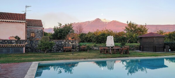 A swimming pool with a stone wall and a wooden deck, a stone building with a tiled roof, and a mountain in the background.