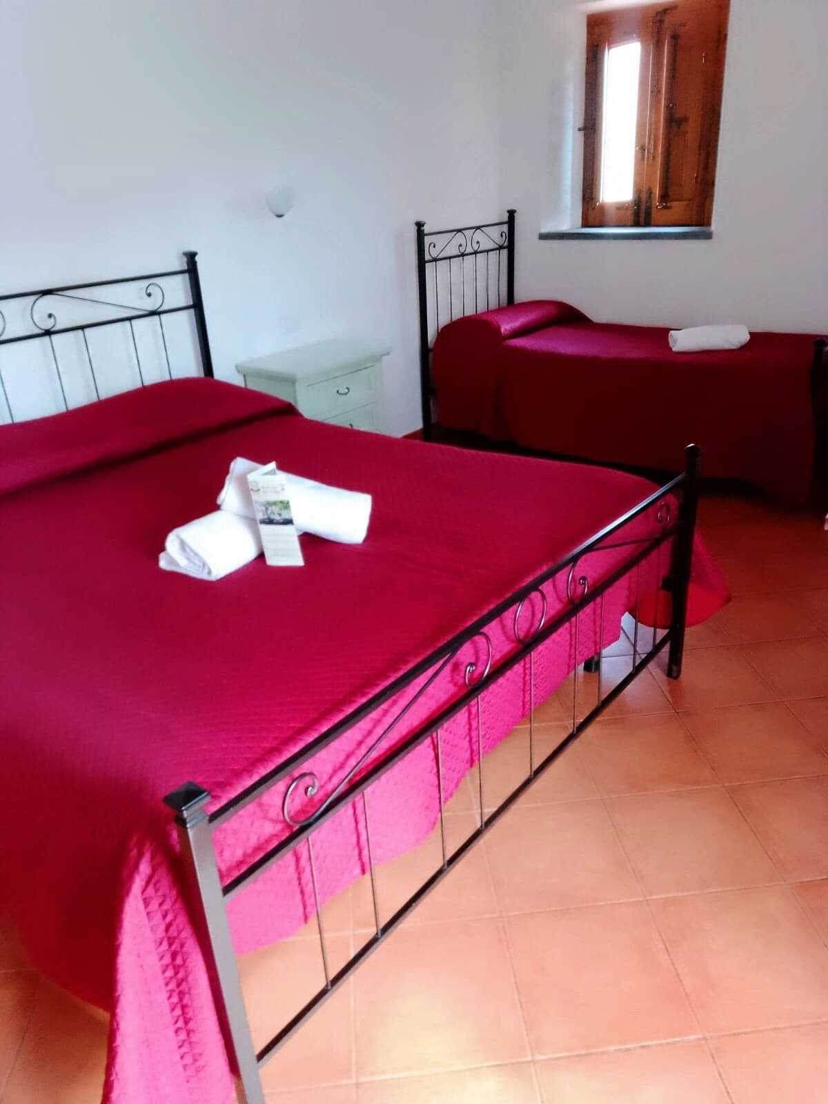 Una stanza con un letto grande, un letto più piccolo, un comodino, una finestra e una parete rossa.