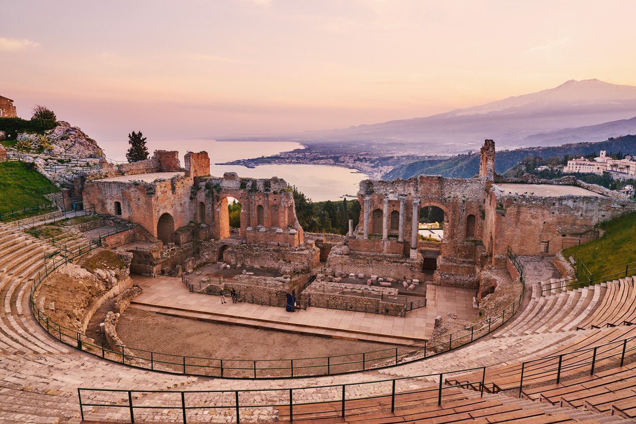 An ancient amphitheater with stone seating and ruins, overlooking a body of water and distant mountains.