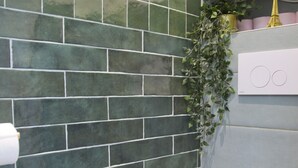 Un baño con inodoro blanco, paredes con azulejos verdes, una planta colgante y un mueble blanco con una estatua dorada.