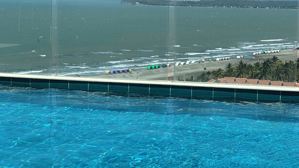 Ein Pool mit klarem, blauem Wasser, ein Strand mit bunten Sonnenschirmen und Blick auf das Meer und das entfernte Land.