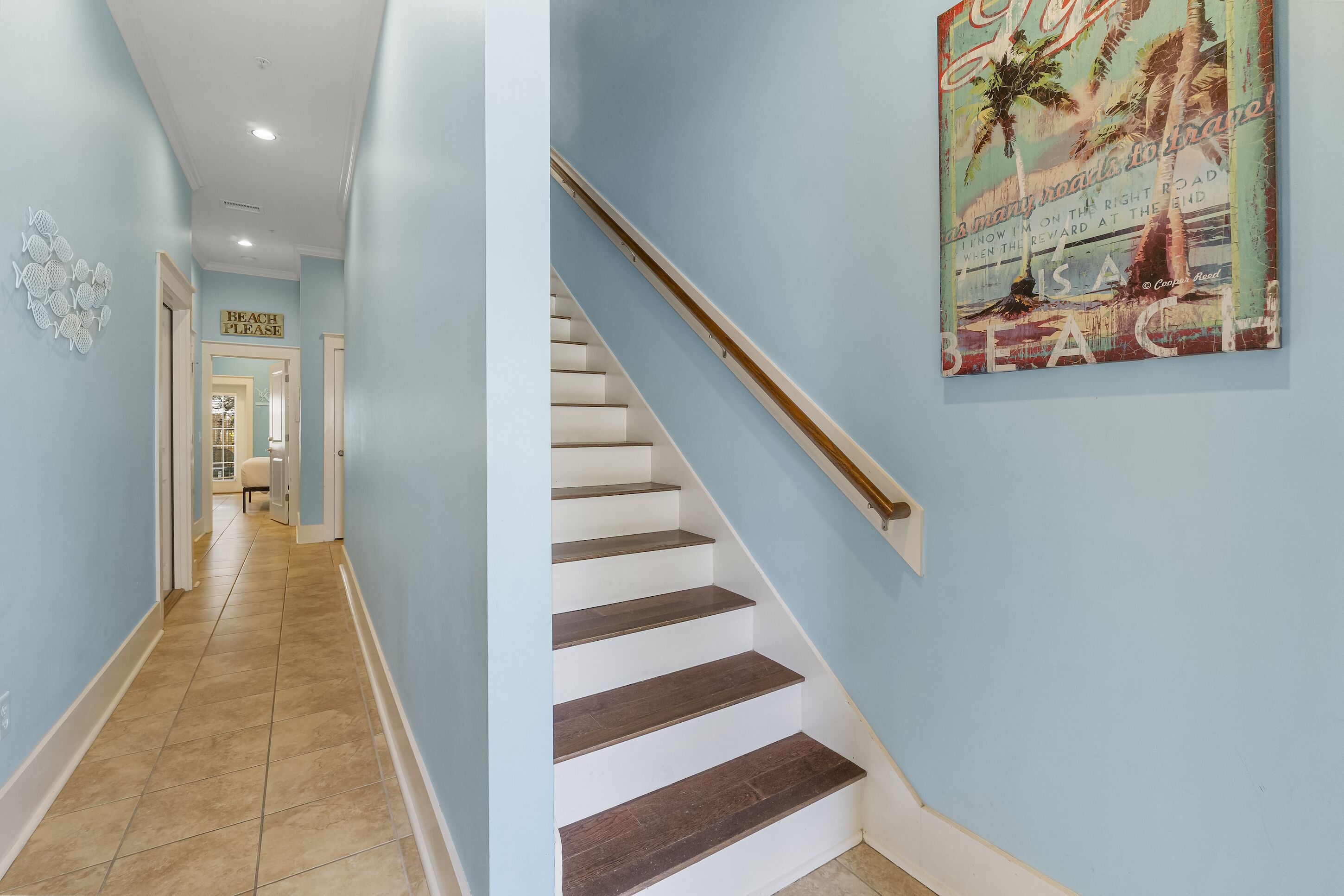 A staircase with white steps and a wooden handrail, leading to a room with light blue walls and a beach-themed poster.