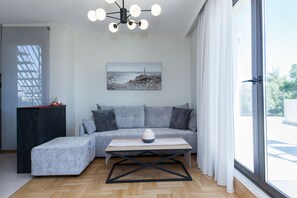A modern living room with a sectional sofa, a wooden coffee table, a painting of a coastal scene, and a large window with curtains. - Marousi Modern Green Oasis with Balcony (Marousi)