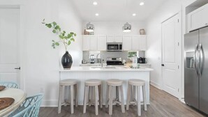 A modern kitchen with white cabinetry, a stainless steel refrigerator, and a bar area with stools. - Experience Seajoy! 200 yard walk to beach! (Orange Beach)