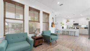 A spacious living room with teal sofas, a wooden side table, and a large window with blinds. A kitchen with white cabinets and a central island is visible in the background. - Experience Seajoy! 200 yard walk to beach! (Orange Beach)