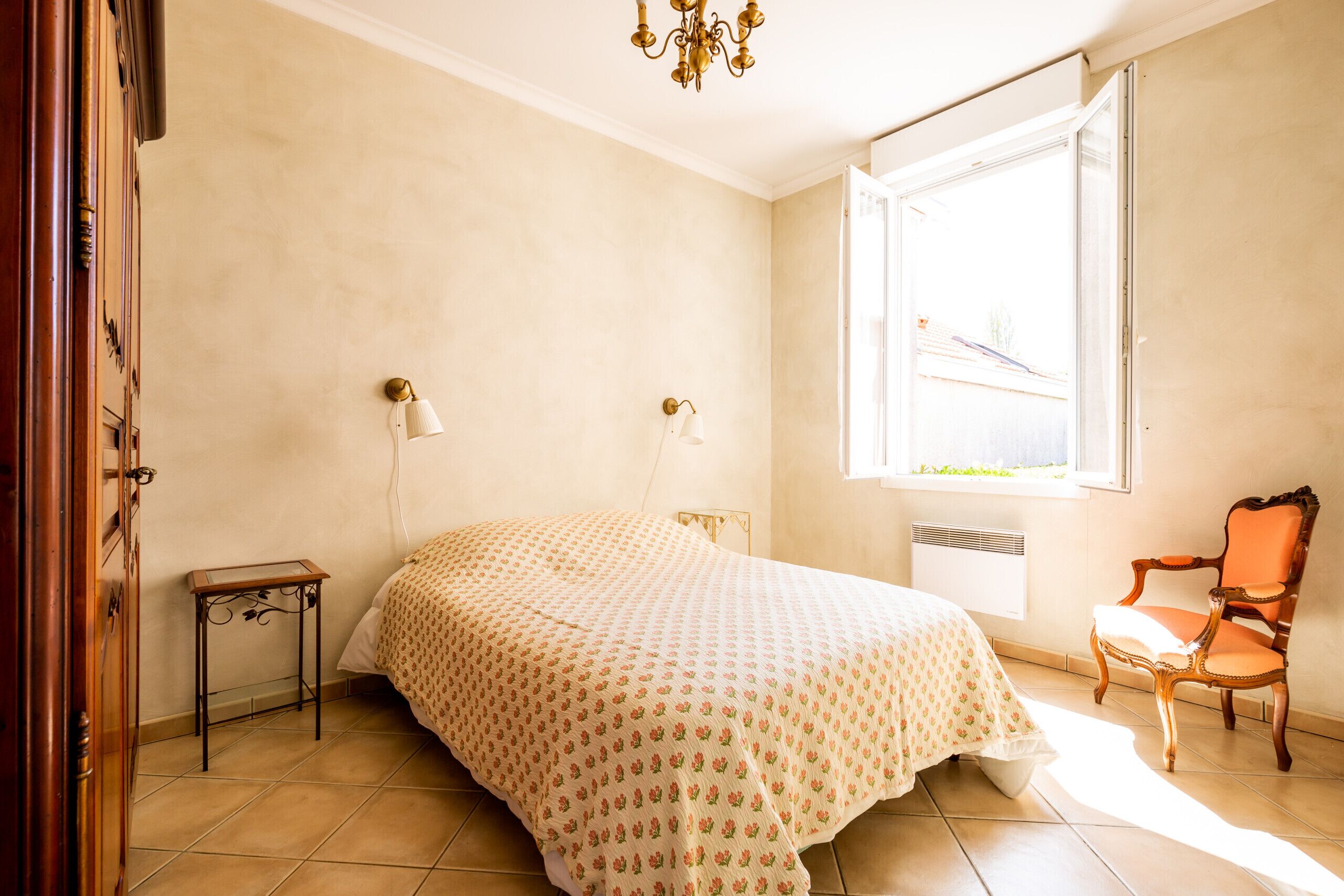 A bedroom with a bed, two bedside lamps, a radiator, a window, and a chair.