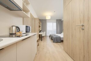 Habitació d'hotel petita amb cuineta, un llit amb llençols grisos i una taula amb cadira.