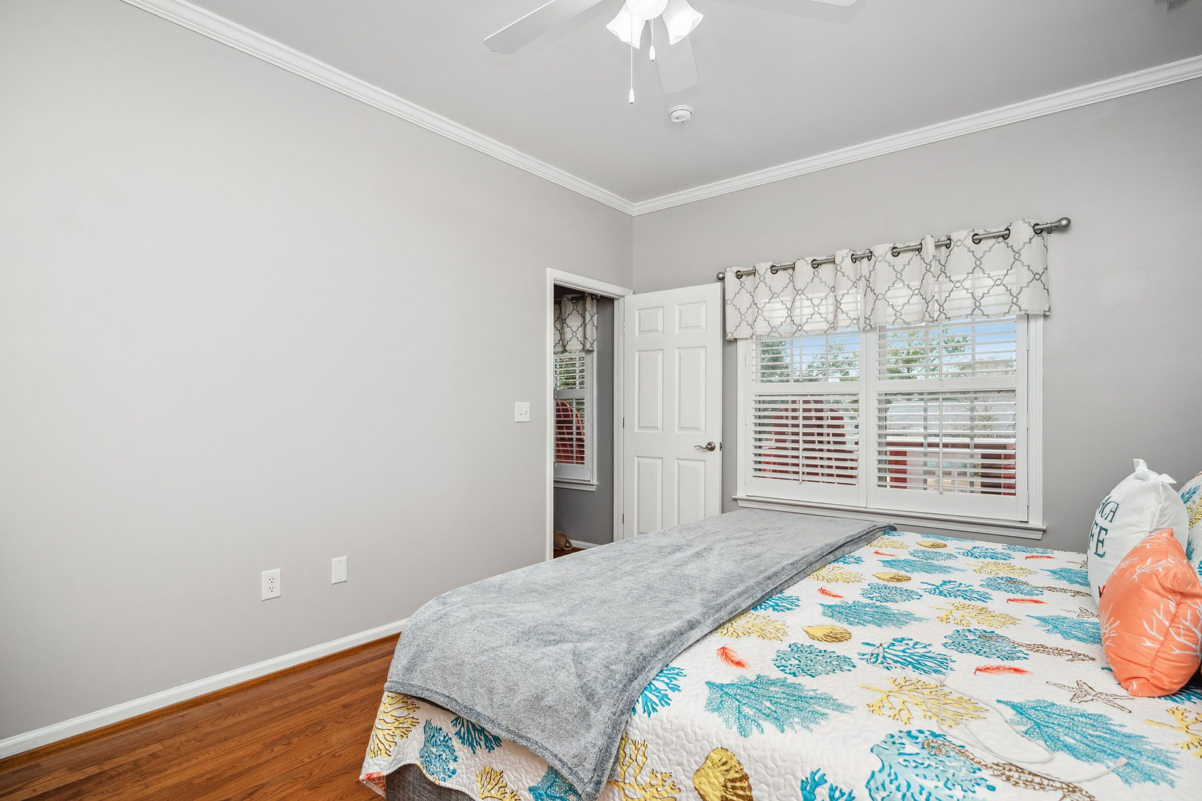 Un dormitorio con cama, ventana con persianas, ventilador de techo y puerta que da a otra habitación.