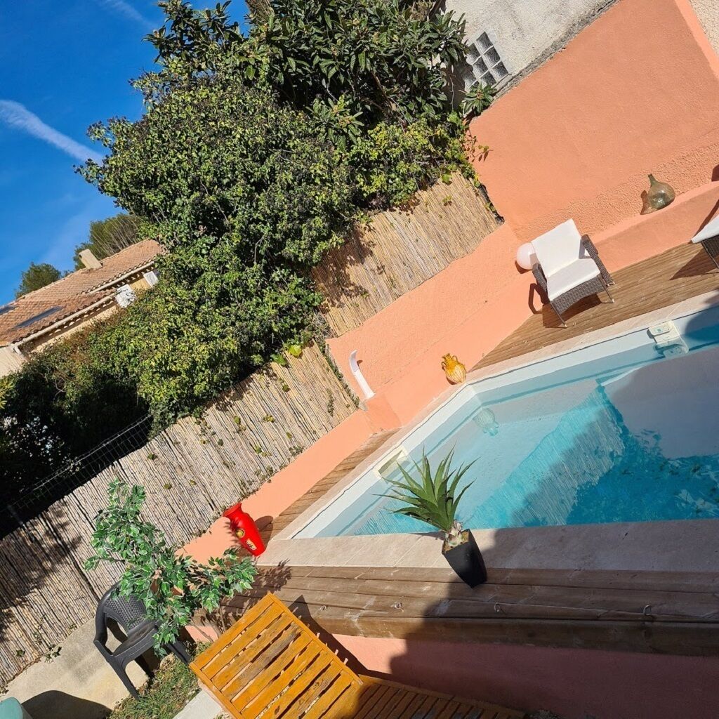 A swimming pool with a wooden deck, surrounded by a fence and potted plants.