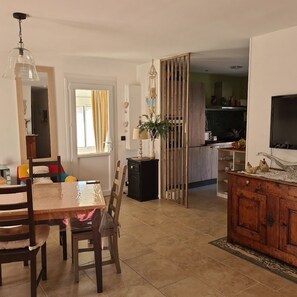 A dining area with a wooden table and chairs, a black cabinet, a TV, and a kitchen in the background.