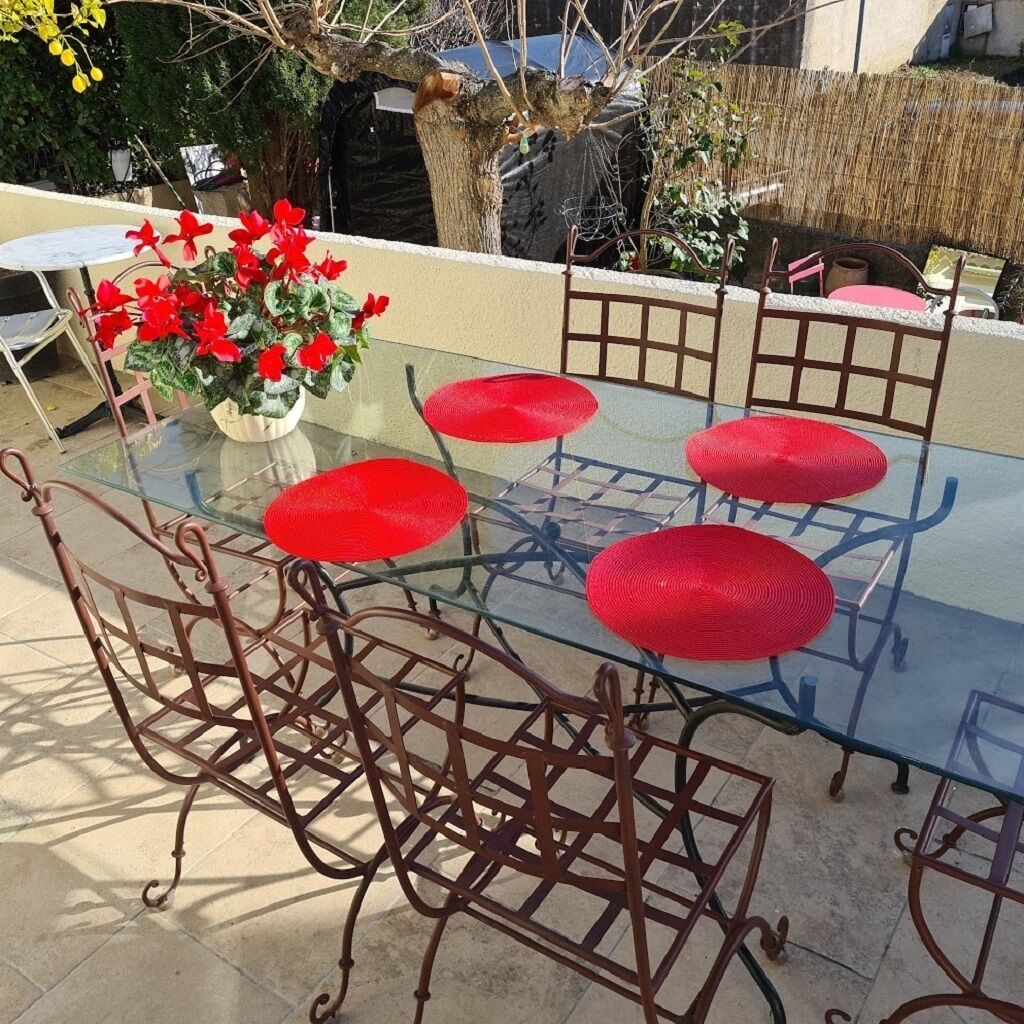 Une table à plateau en verre, entourée de tabourets rouges en osier et de chaises en métal, dans un décor extérieur agrémenté d’un jardin et d’un arbre en arrière-plan.