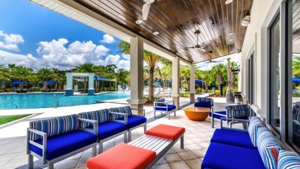 A covered outdoor seating area with blue and red cushioned sofas, a pool, and a view of the pool and greenery.