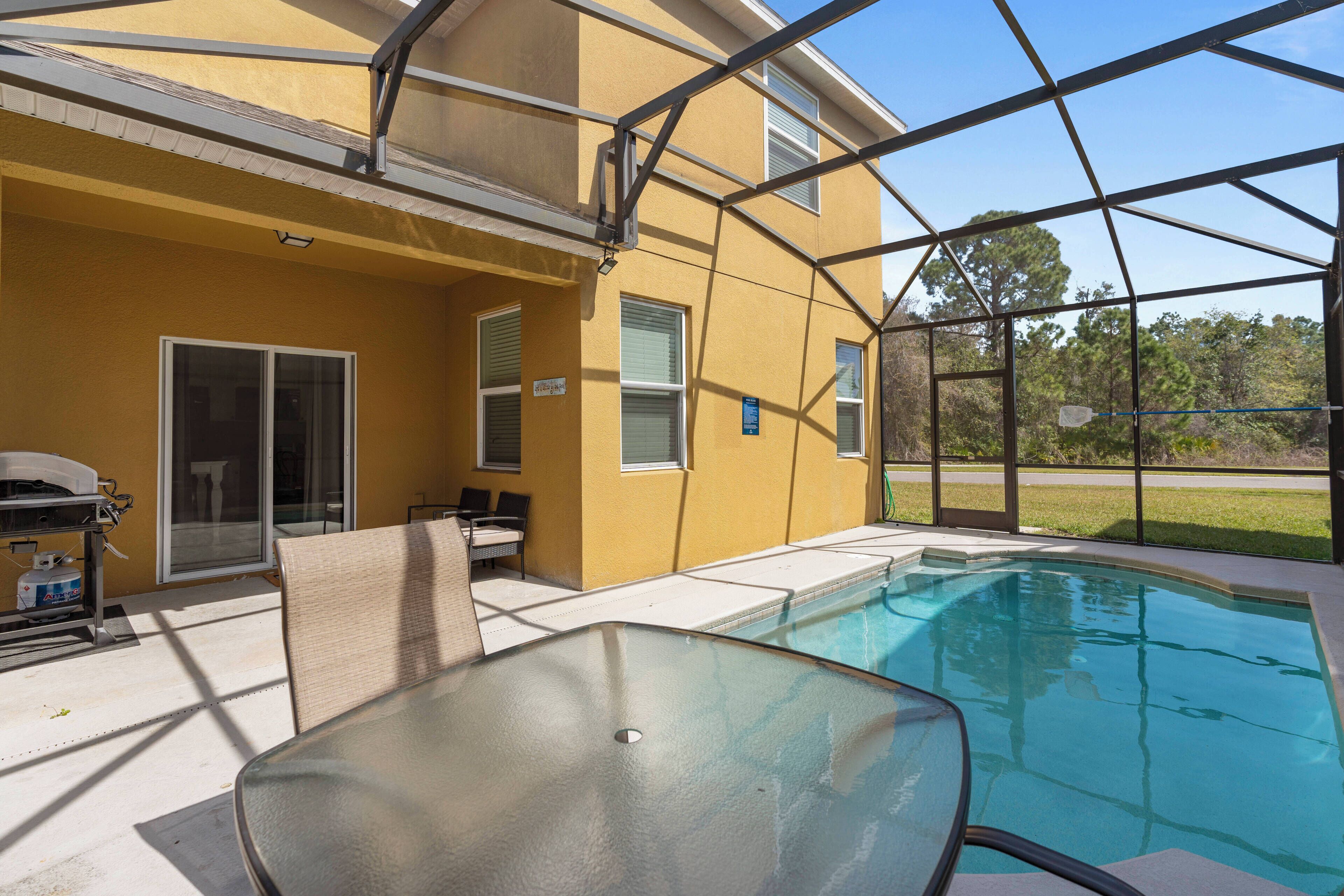 A covered outdoor pool area with a glass table and chairs, a barbecue grill, and a yellow building with windows.