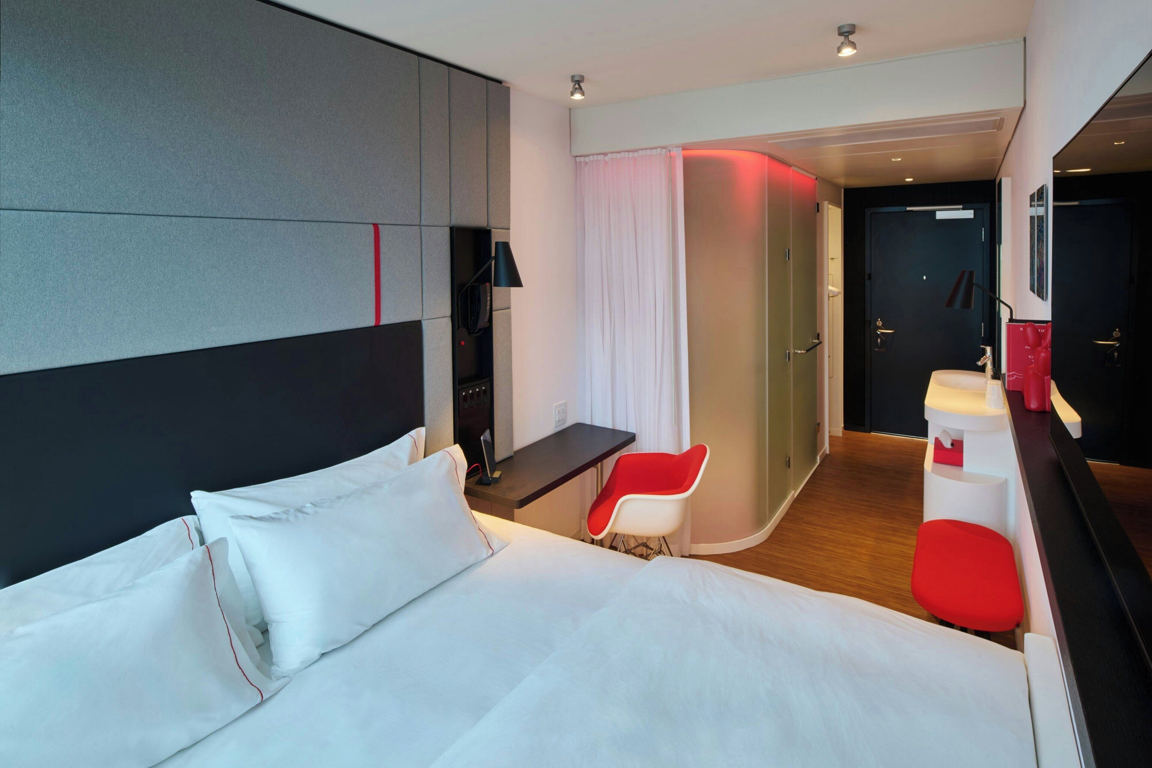 A modern hotel room with a large bed, a desk, a red chair, and a small red stool.