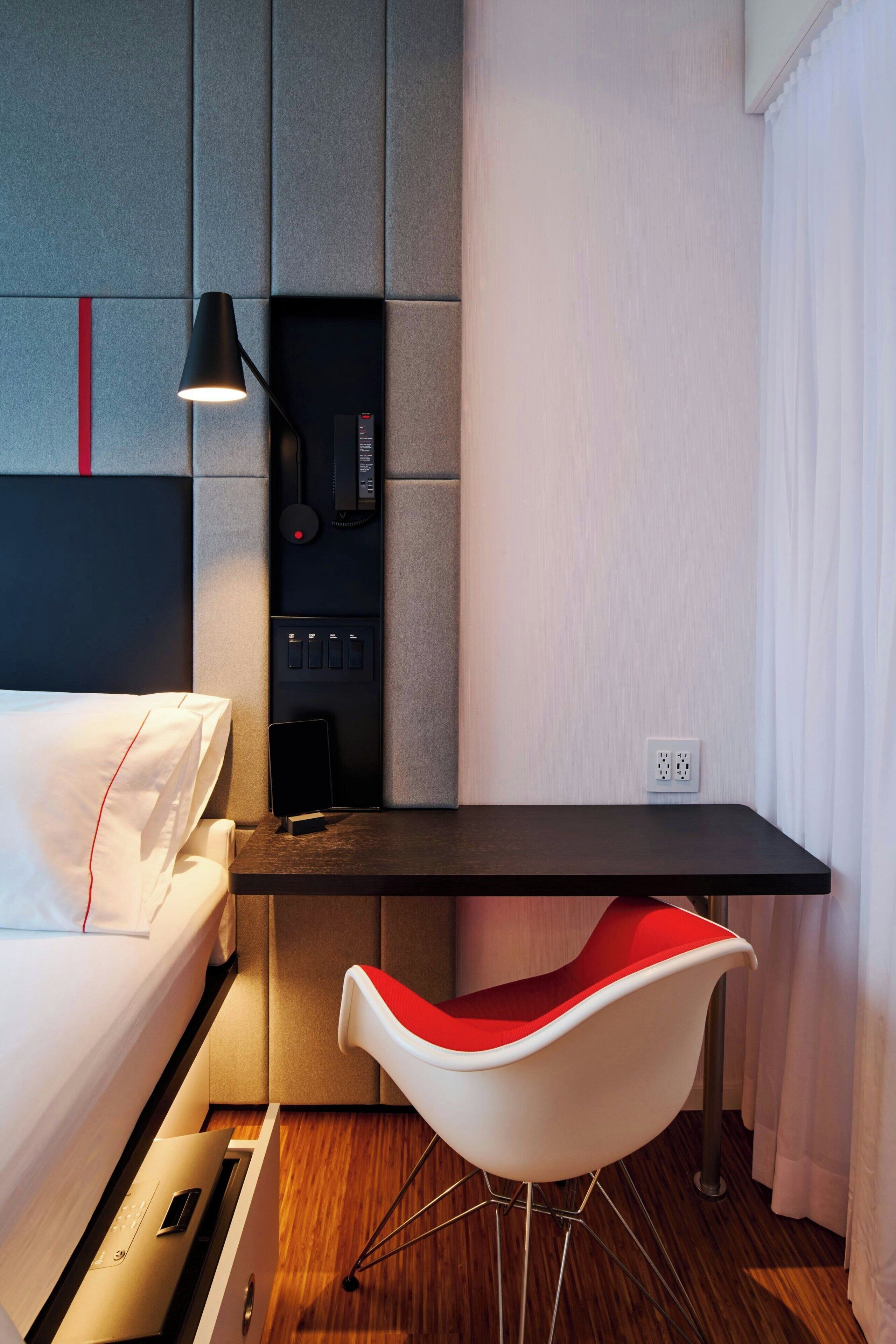 A compact hotel room with a bed, a desk, a modern chair, a lamp, and a phone on the wall.