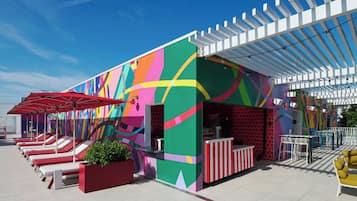 A colorful outdoor seating area with red and white striped tables and chairs, a covered section with a white pergola, and a vibrant mural on the building's exterior.