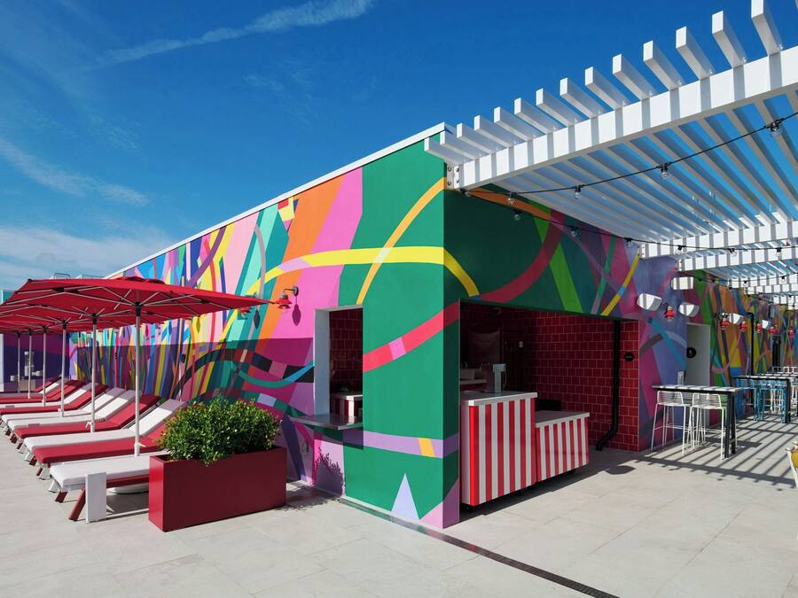A colorful outdoor seating area with red and white striped tables and chairs, a covered section with a white pergola, and a vibrant mural on the building's exterior.