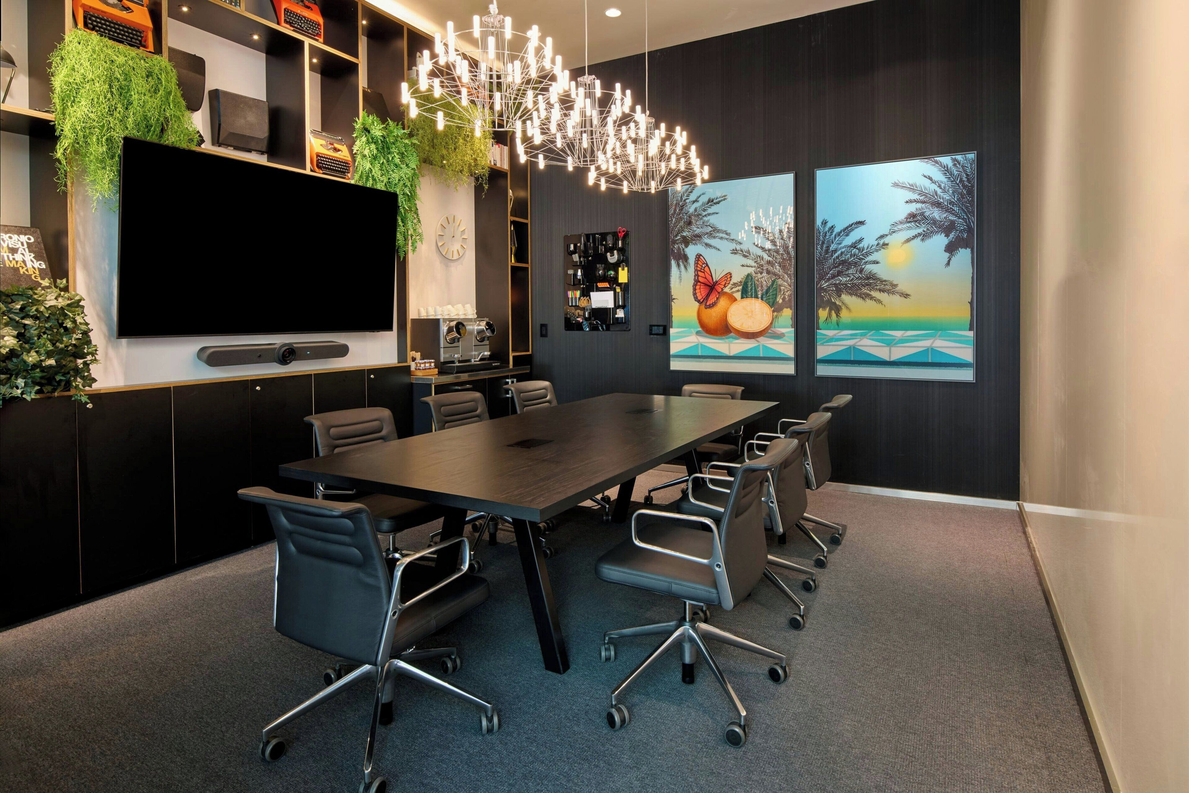A modern meeting room with a large table, chairs, a flat-screen TV, and decorative plants.
