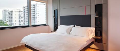 Una habitación de hotel moderna con una cama grande, un cabecero integrado, mesitas de noche y vistas a edificios altos a través de un ventanal.