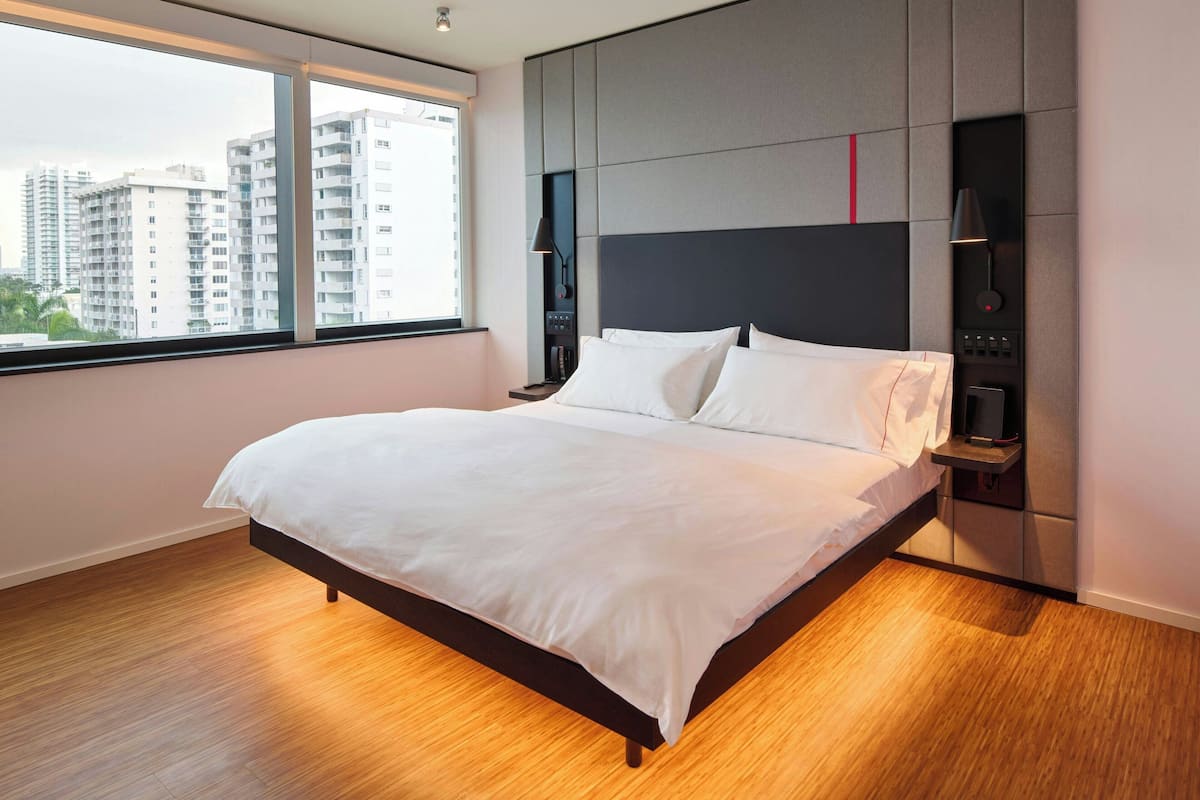 A modern hotel room with a large bed, a nightstand, a wall-mounted control panel, and a window with a city view.