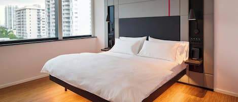 A modern hotel room with a large bed, a nightstand, a wall-mounted control panel, and a window with a city view.