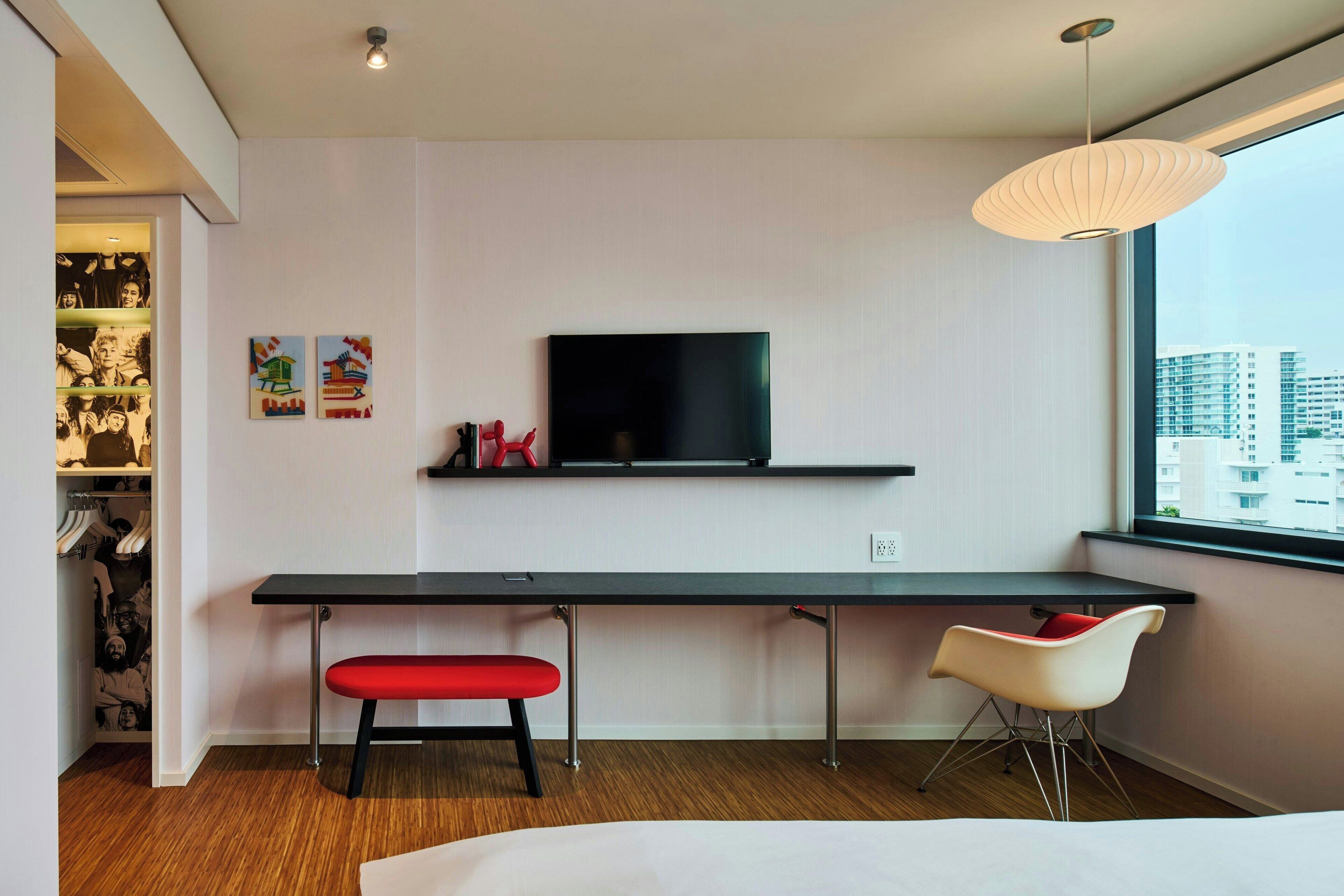 A modern hotel room with a large bed, a desk with a chair, a TV mounted on the wall, and a window with a city view.