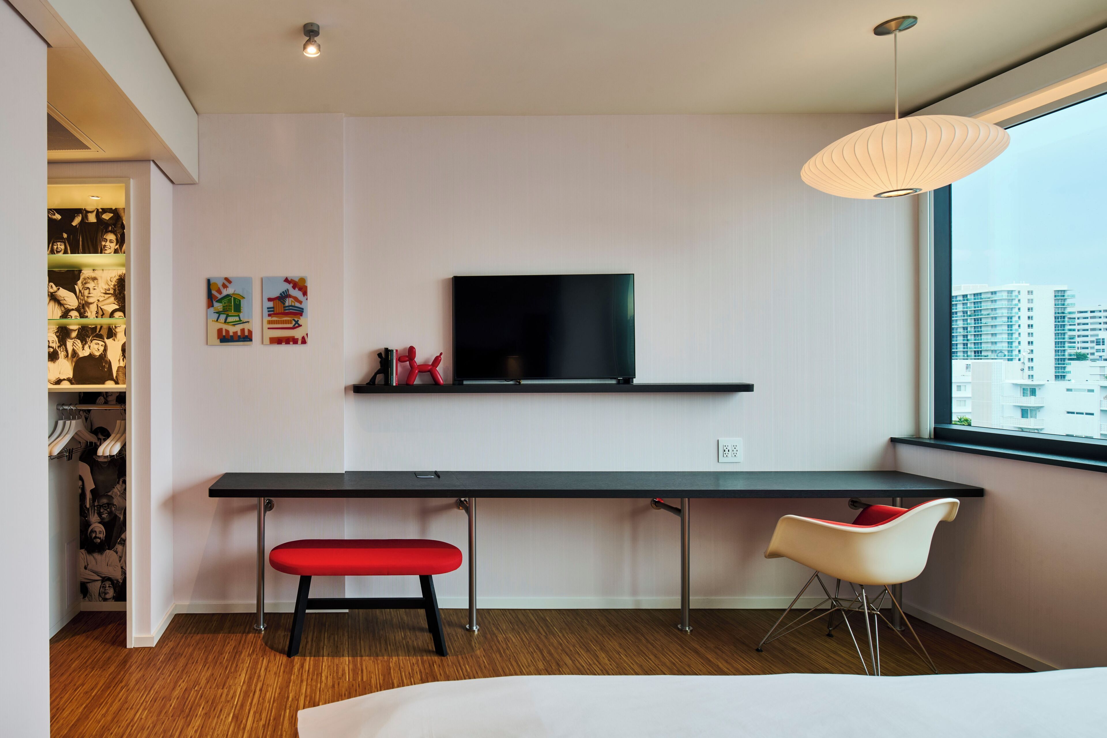 Una habitación moderna con un escritorio amplio, un banco rojo, un televisor y una ventana con vistas a la ciudad.