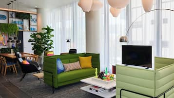 A modern living room with a green sofa, a coffee table, and a TV. There is a dining area with wooden furniture and a plant.