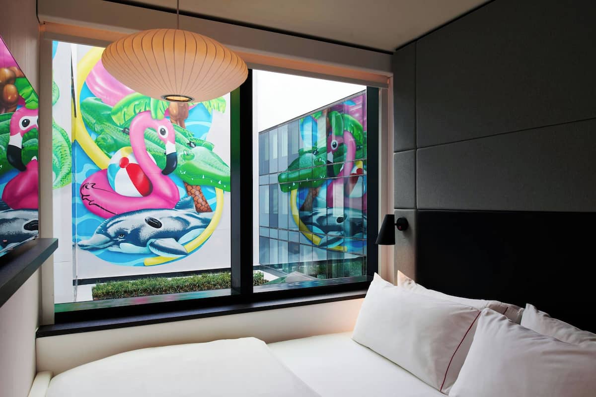 A modern hotel room with a large window, a bed, and a wall mural featuring colorful animals.