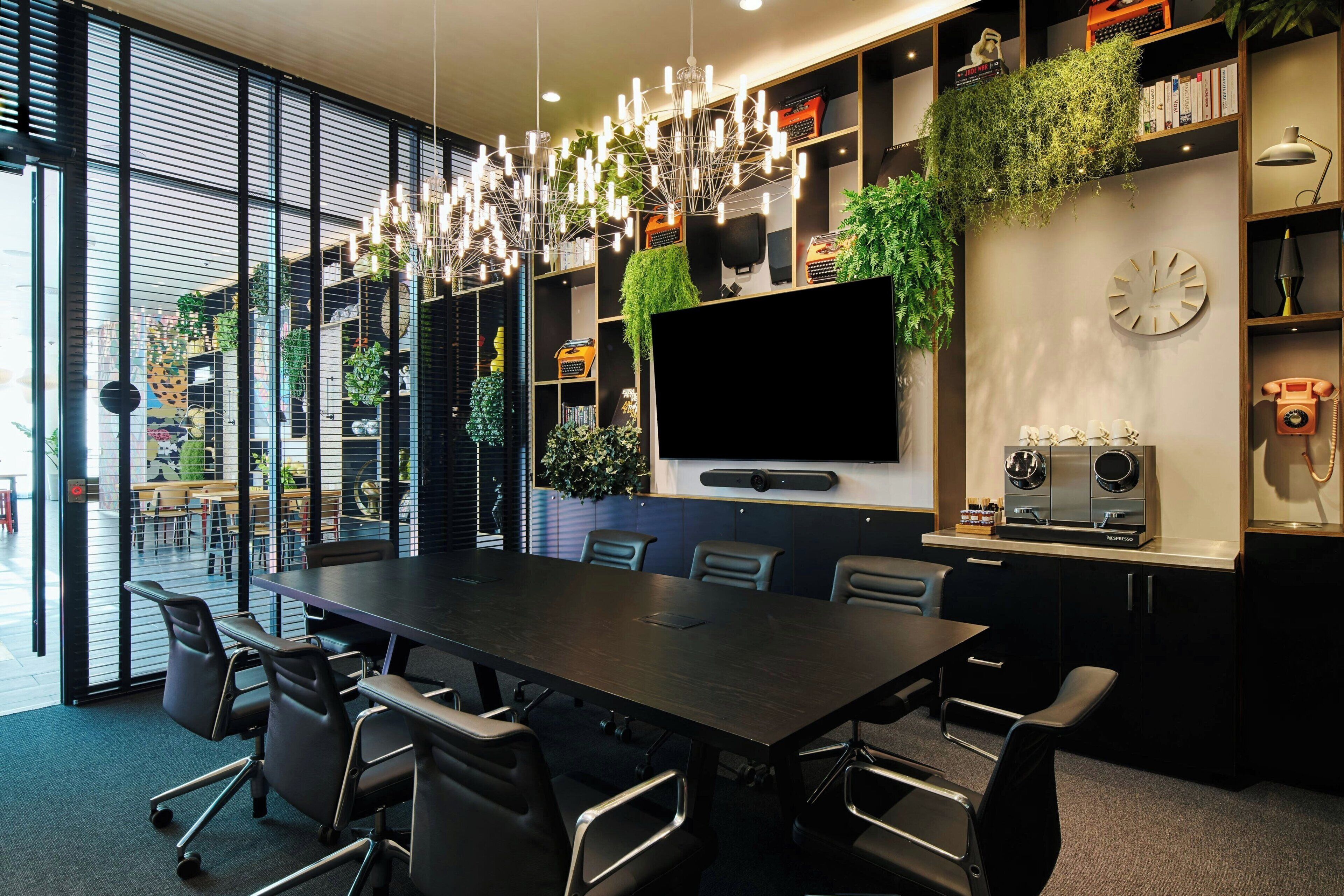 A modern conference room with a long table, chairs, a large screen, and shelves with plants and books.