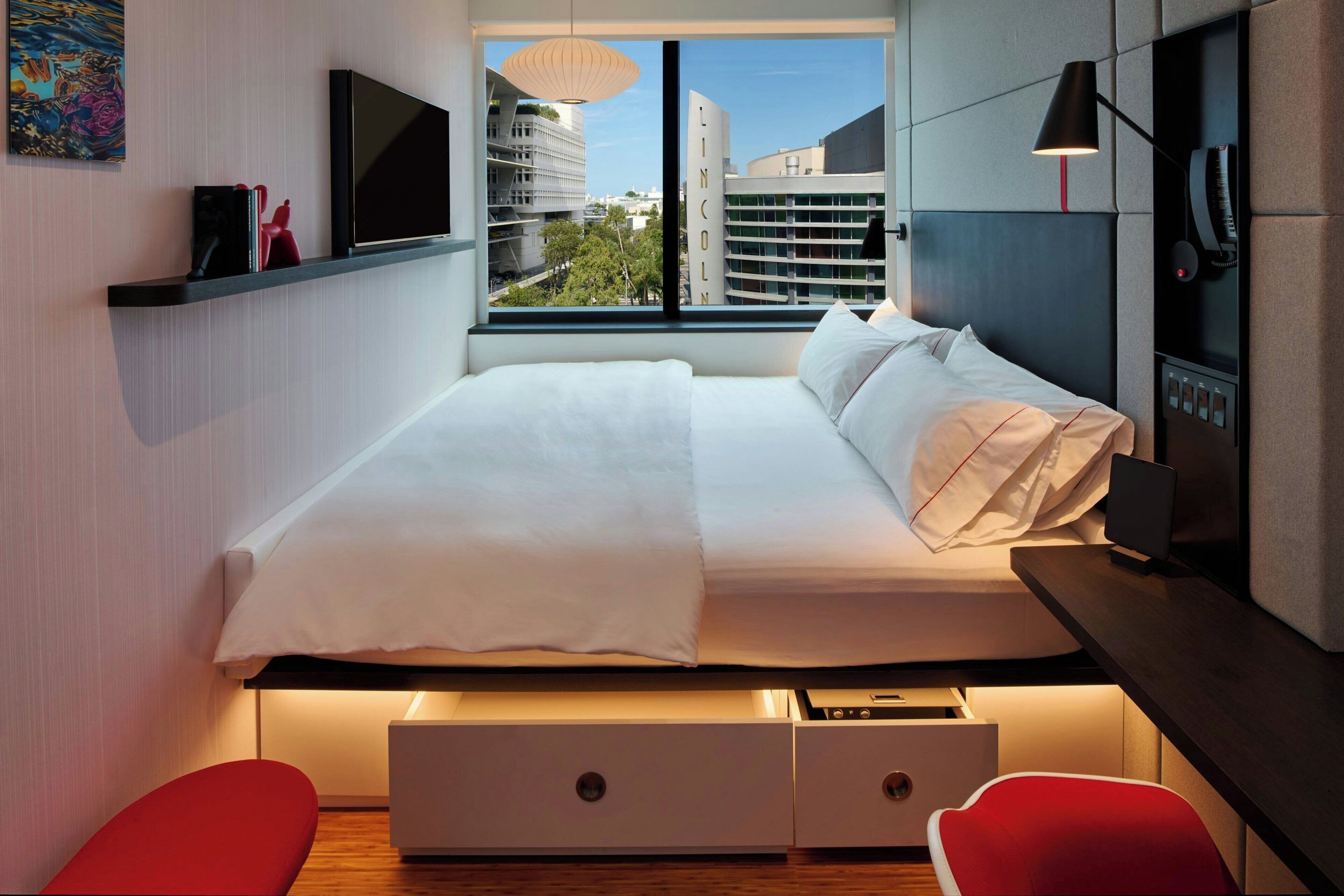 A modern hotel room with a bed, a TV, a desk, and a window with a city view.