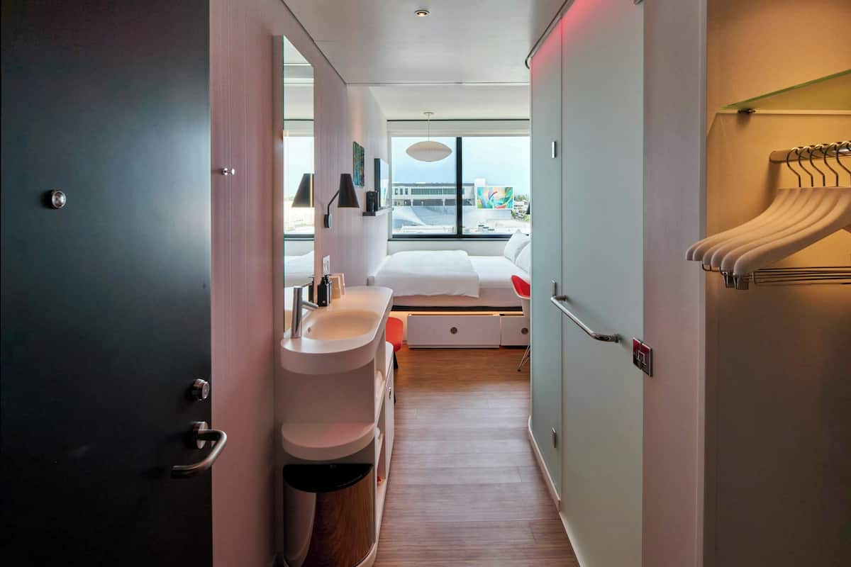 A modern hotel room with a large bed, a bathroom with a sink and mirror, and a view of the city through large windows.