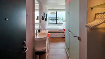 Una habitación de hotel moderna con una cama grande, un baño con lavamanos y espejo, y vistas a la ciudad a través de amplios ventanales.