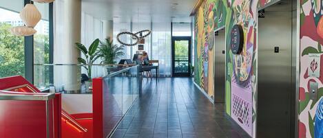 A modern hotel lobby with a glass railing, a red seating area, and an elevator with colorful wall art.