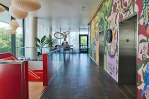 A modern hotel lobby with a glass railing, a red seating area, and an elevator with colorful wall art.