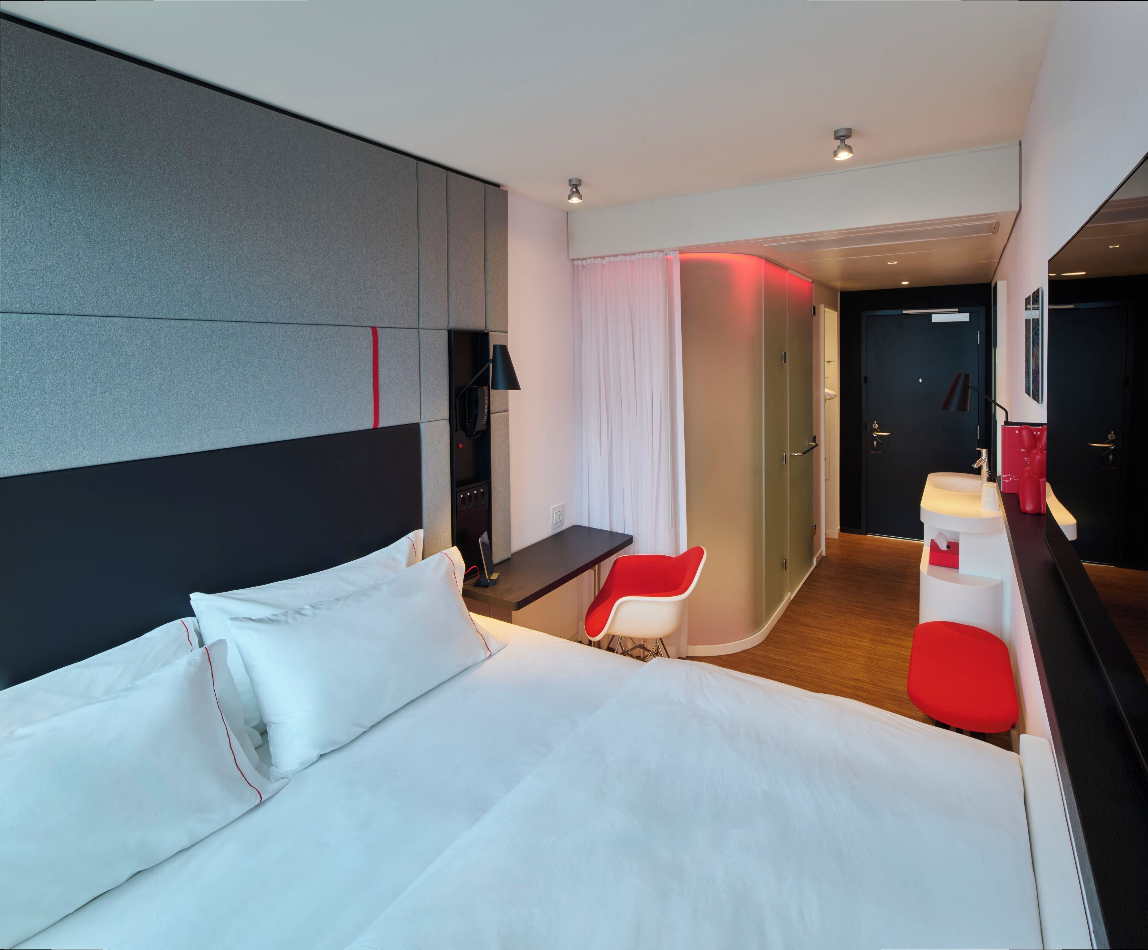 A modern hotel room with a large bed, a desk, a chair, and a bathroom with a mirror and sink.