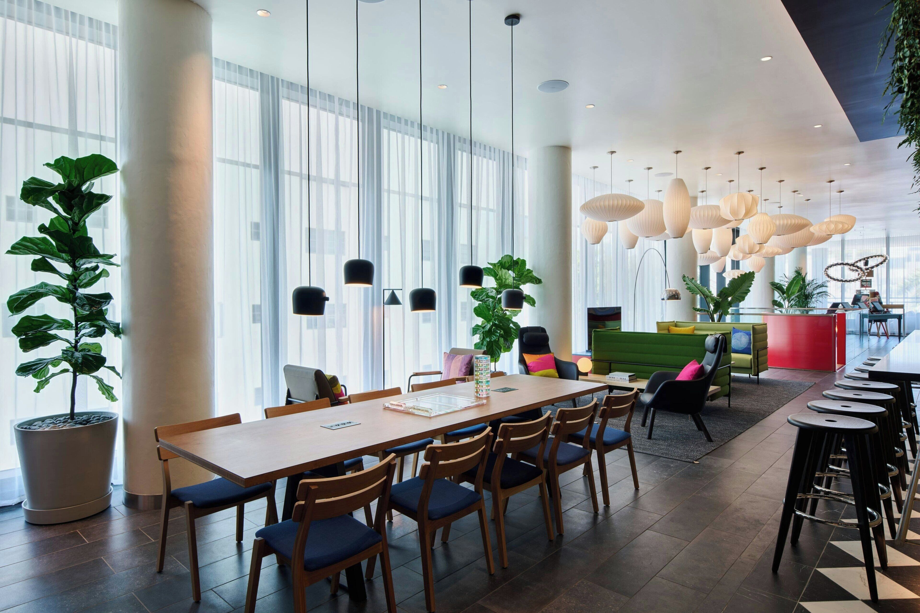 A modern hotel lobby with a long wooden table, multiple chairs, and hanging pendant lights. There are large windows with sheer curtains, potted plants, and a reception desk in the background.