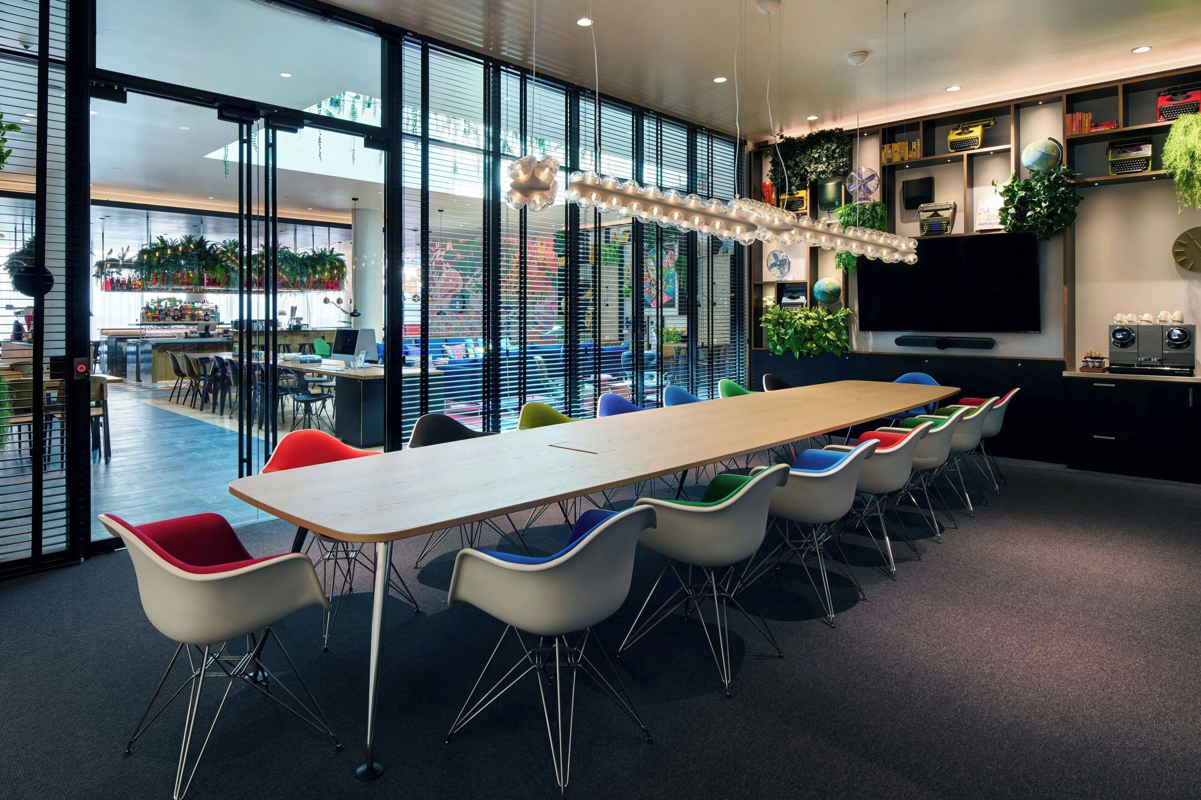 A modern meeting room with a long table, colorful chairs, a large window, and a television.