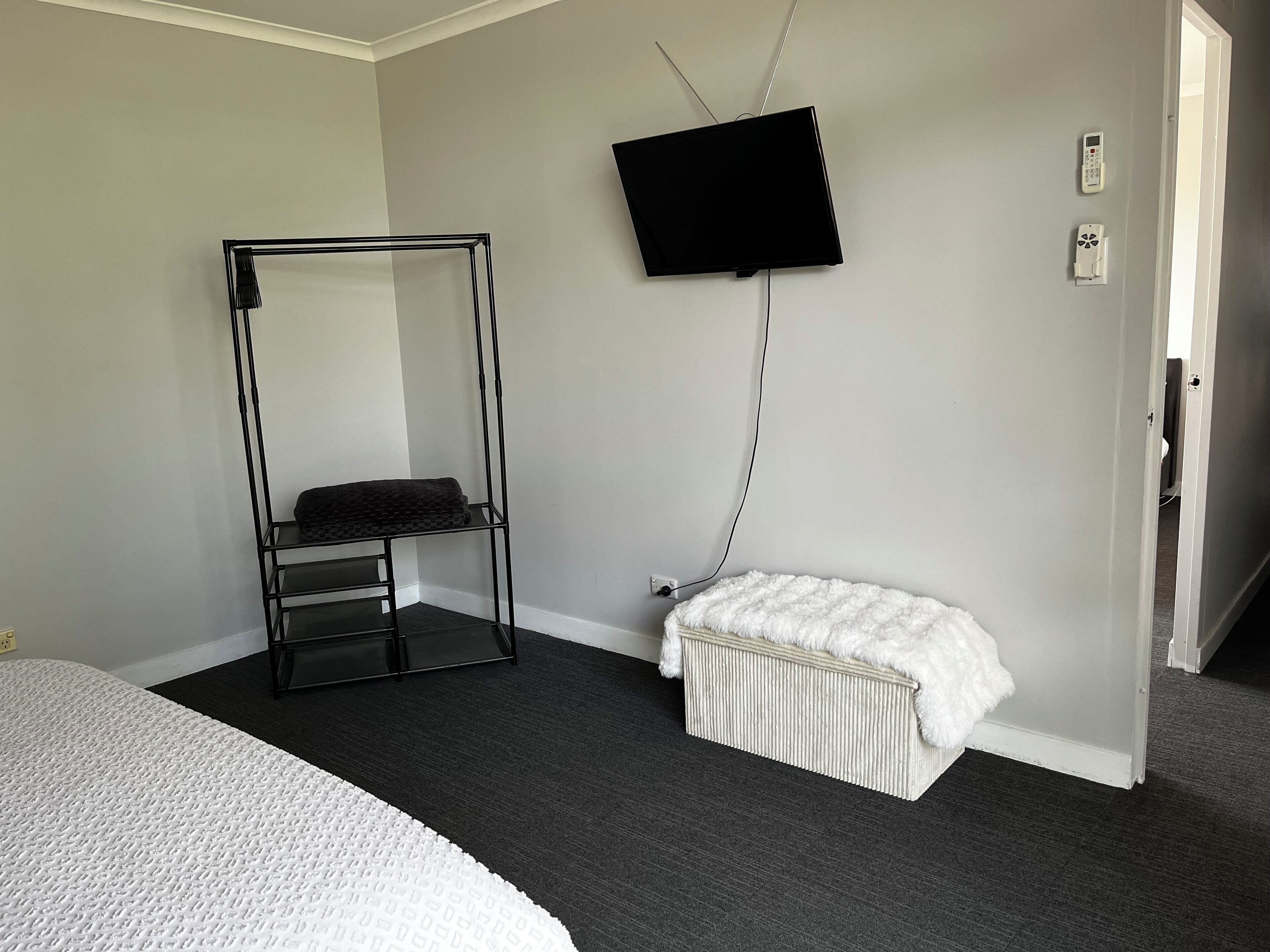 Ein Schlafzimmer mit einem Bett, einem schwarzen Metallregal, einer weißen, flauschigen Bank, einem an der Wand montierten Fernseher und einer Tür zu einem anderen Raum.